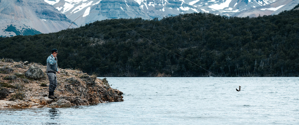 Man fly fishing with fish jumping on the end of his line with mountains in the background